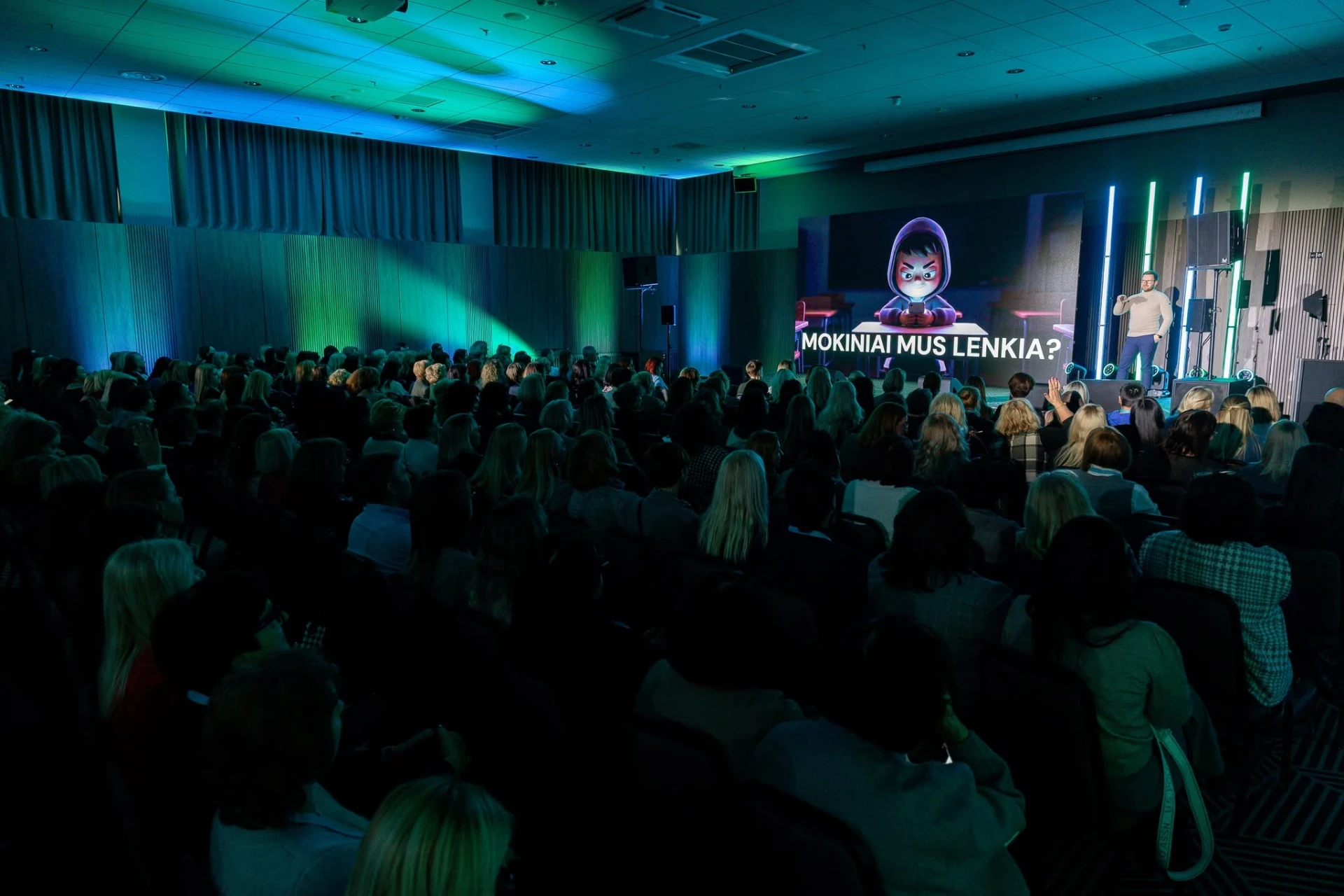 Speaker presenting on a neon-lit conference stage in front of a large audience